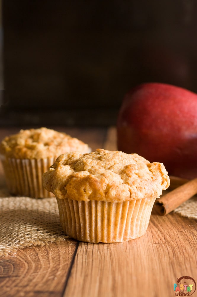 two streusel top apple muffins on wooden table with an apple and cinnamon sticks