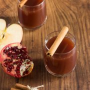 two glasses pomegranate and apple mocktail with cinnamon sticks, a sliced apple and pomegranate