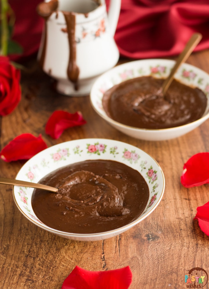 two bowls of chocolate soup with a spoon in each bowl surrounded by rose petals and a porcelain jug dripping with soup and a red satin tablecloth and a red rose in the background