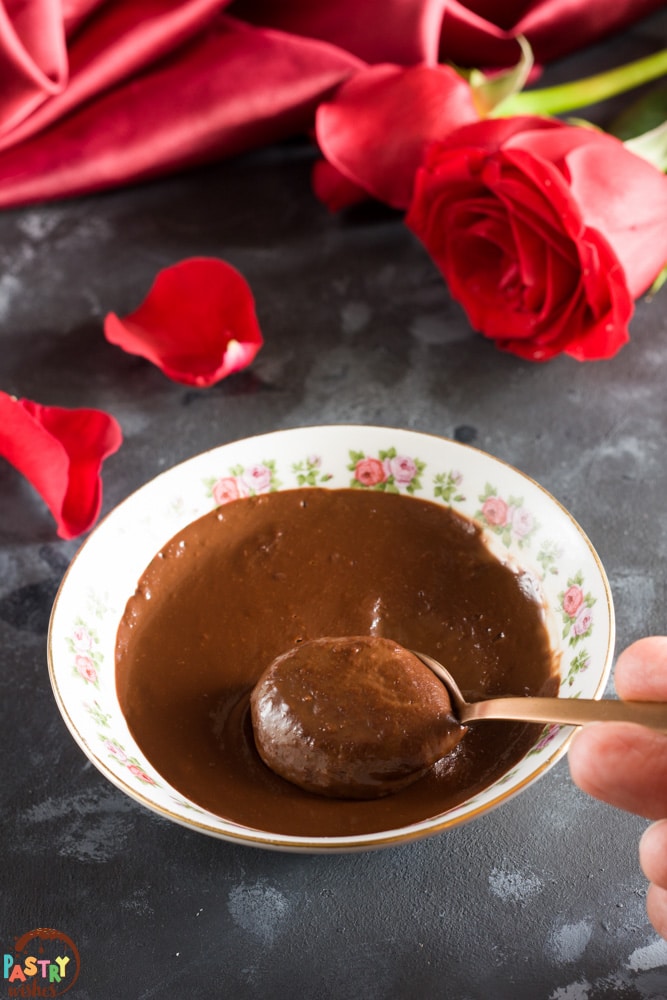 a bowl of chocolate soup with a hand holding a spoon in it and a red rose and red satin tablecloth in the background