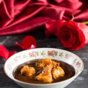 a bowl of chocolate soup with caramel brioche croutons on top and a red rose and red satin tablecloth in the background
