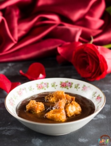 a bowl of chocolate soup with caramel brioche croutons on top and a red rose and red satin tablecloth in the background