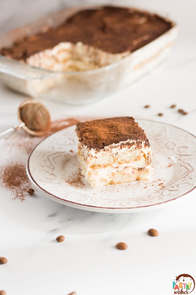 tiramisu without raw eggs on a plate on a white countertop with a pan of tiramisu in the background