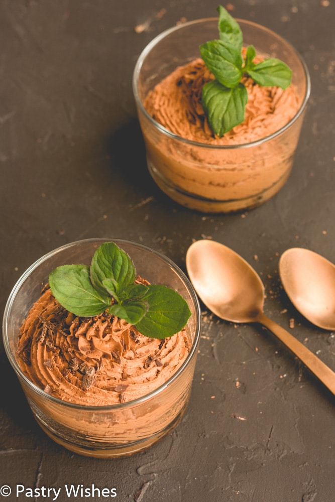 two ingredient chocolate mousse in a glass with mint leaves and chocolate shavings on top on a dark surface with 2 spoons and another glass of mousse in the background