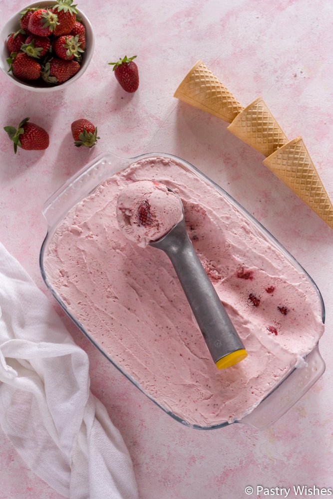 strawberry ice cream with a scoop on a pink and white surface with strawberries and cones