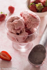 a bowl of strawberry ice cream with a scoop and a bowl of strawberries on a pink and white surface