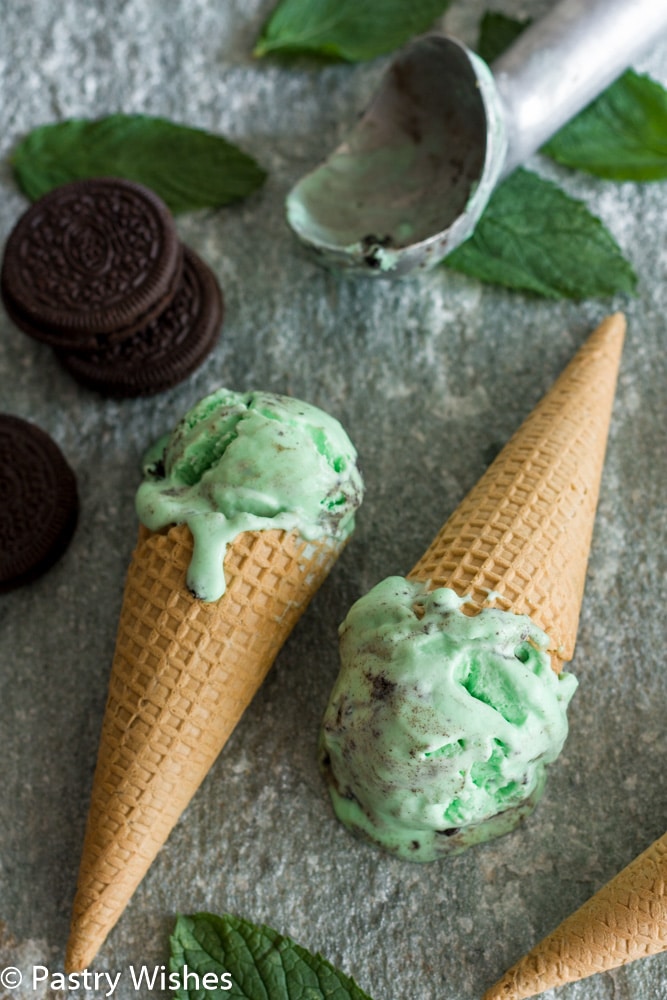 2 cones with oreo mint ice cream on a gray surface with oreo cookies and a scoop in backbround
