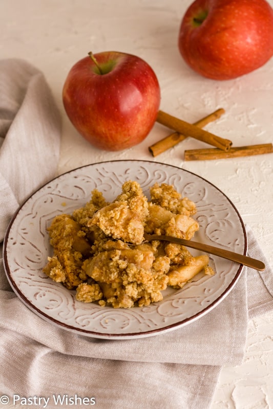 vegan apple crisp on a plate with apples and cinnamon sticks in the background