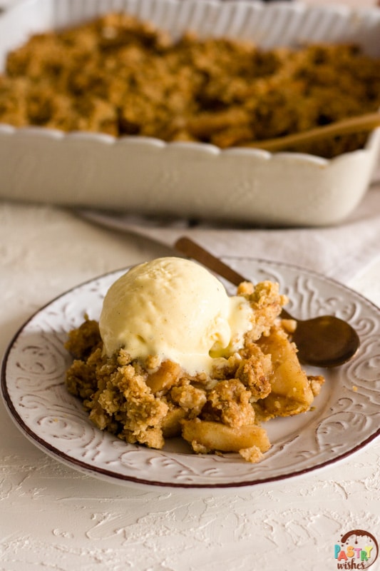 vegan apple crisp on a plate topped with ice cream and a baking dish in the background