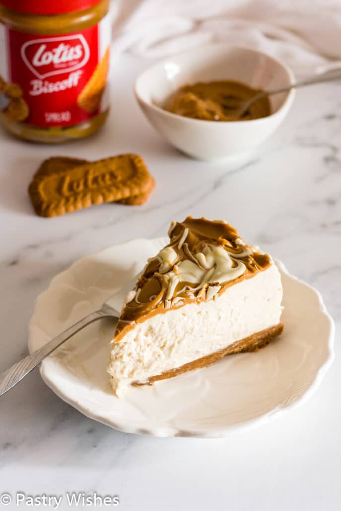 a-slice-of-biscoff-cheesecake-on-a-white-plate-with-biscoff-cookies-and-spread-in-the-background