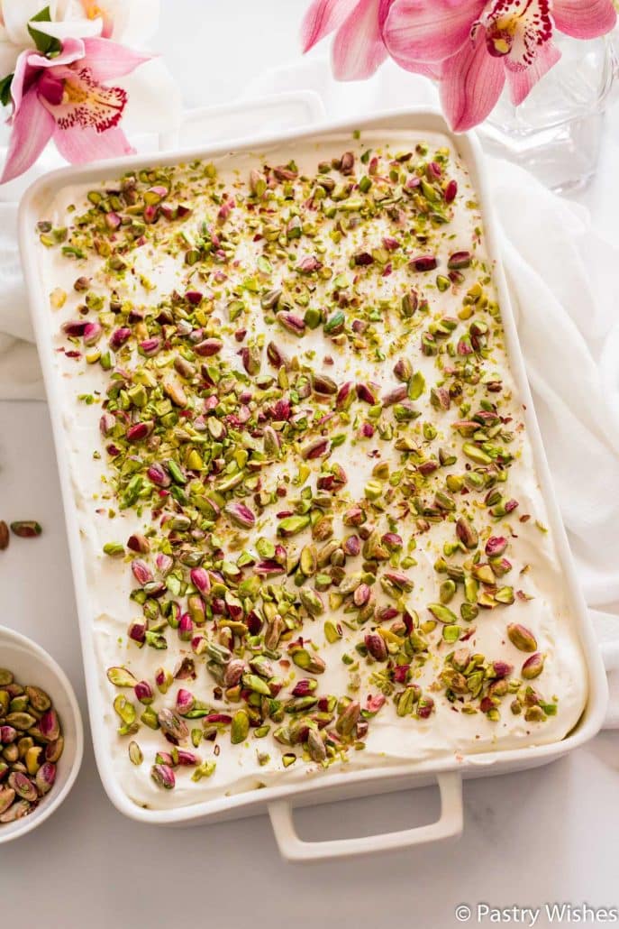 a white baking dish with greek ekmek kataifi topped with pistachios and pink flowers in the background