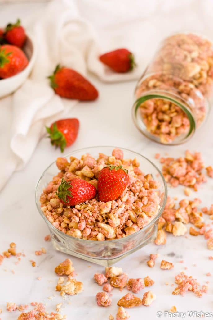 strawberry crunch topping in a jar and a bowl on a white surface with strawberries in background