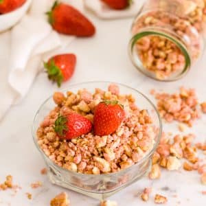 Strawberry crunch topping in a jar and a bowl on a white surface with strawberries in background.
