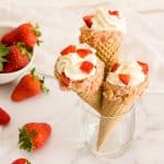 Three strawberry crunch cheesecake cones in a glass with strawberries in the background.