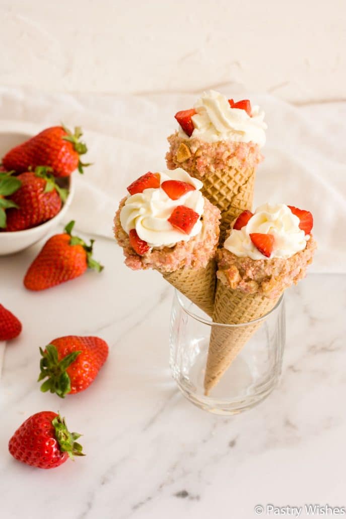 three strawberry crunch cheesecake cones in a glass with strawberries in the background