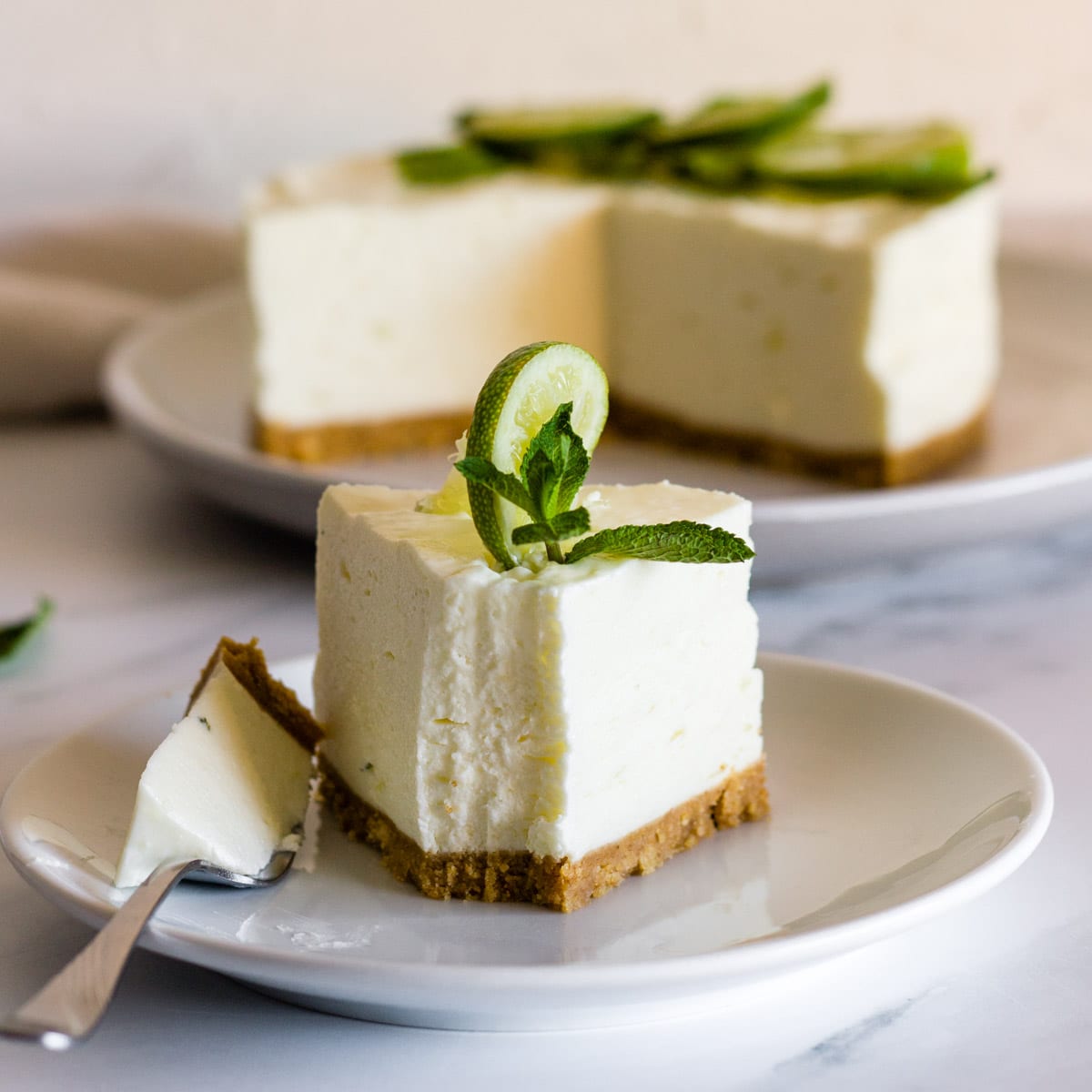 a slice of mojito cheesecake with a slice of lime and a sprig of mint on a white plate with a fork 
