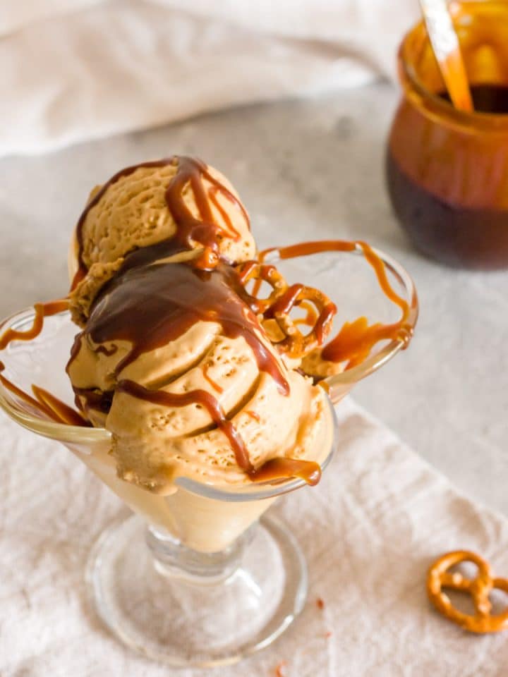 Salted caramel pretzel ice cream in a glass with a jar of caramel in the background.