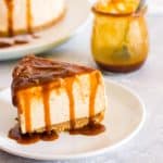 A slice of no bake salted caramel cheesecake on a white plate with cheesecake and a jar of caramel sauce in the background.