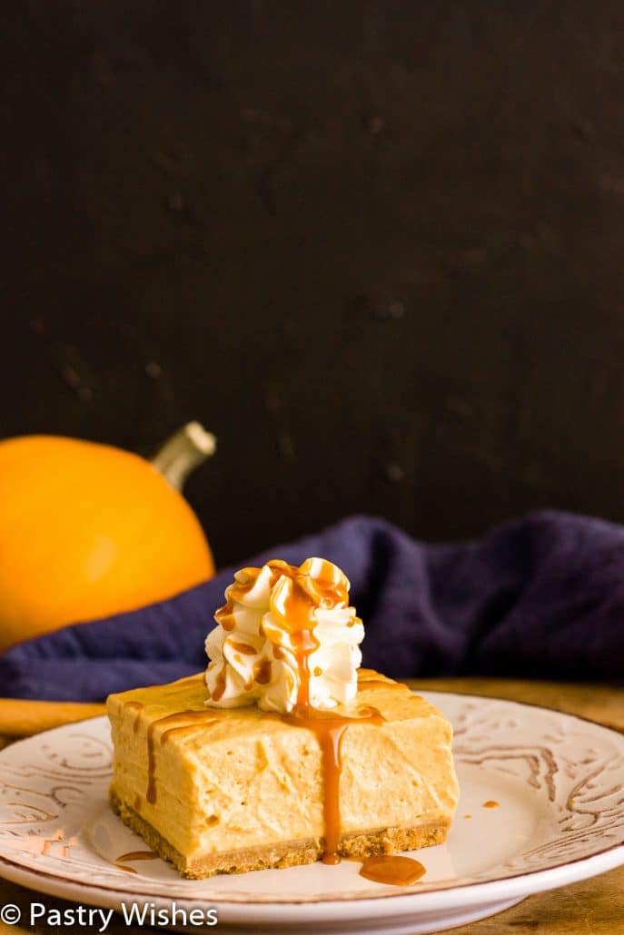 a slice of pumpkin cheesecake bar with whipped cream and caramel sauce on top and pumpkin in back