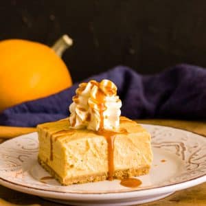 A slice of pumpkin cheesecake bar with whipped cream and caramel sauce on top and a pumpkin in the back.