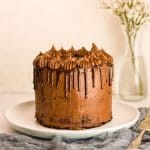 Chocolate drip cake on a white plate with gray napkin and flowers in a vase in background.