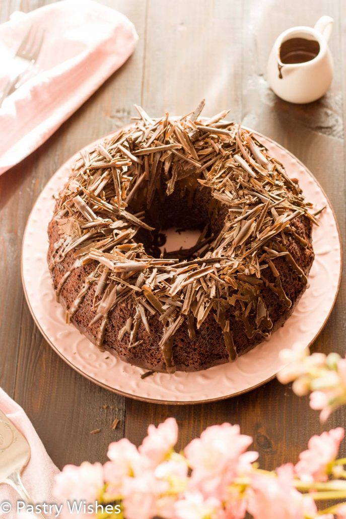 chocolate vegan bundt cake on a pink plate topped with chocolate glaze and chocolate shavings on a wooden table with a pink napkin and flowers