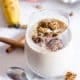 A granola smoothie in a glass topped with granola clusters on a white surface with cinnamon sticks and a banana.
