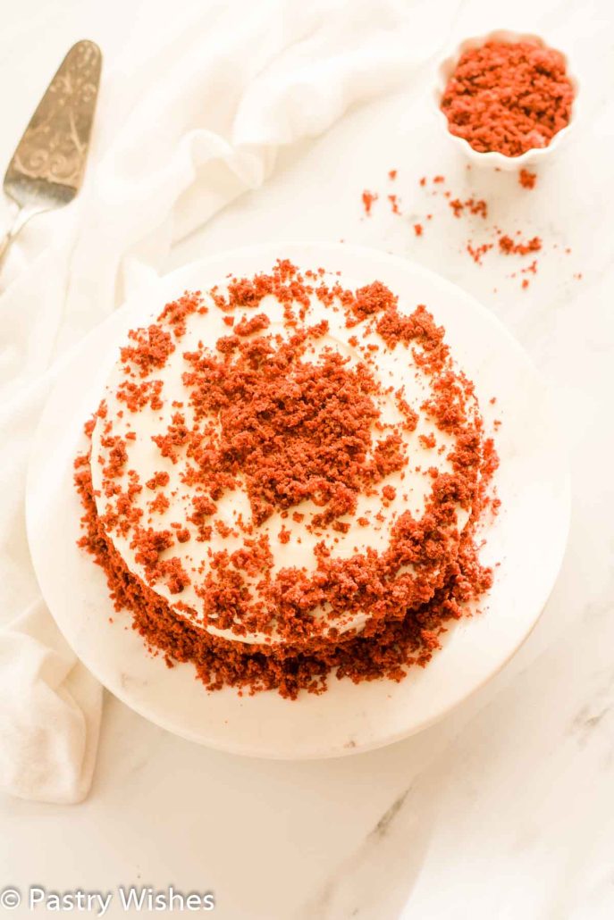 An overhead image of eggless red velvet cake on a white marble cake platter next to a cake server and red cake crumbs.