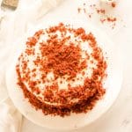 An overhead image of eggless red velvet cake on a white marble cake platter next to a cake server and red cake crumbs.