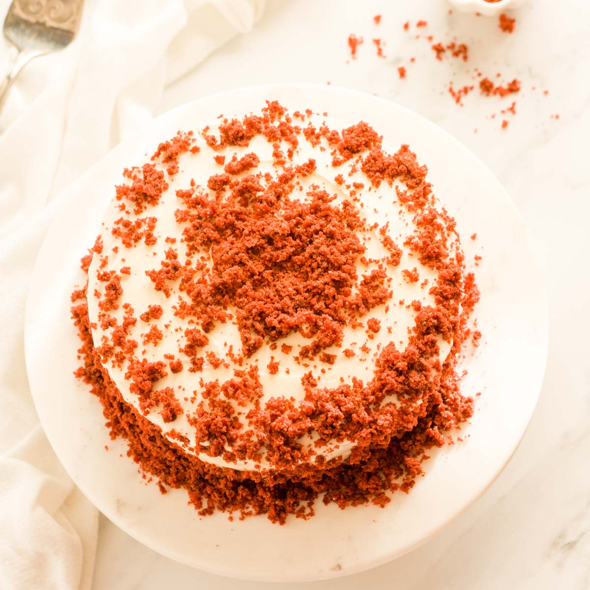 Eggless red velvet cake on a white cake platter with red cake crumbs on top.