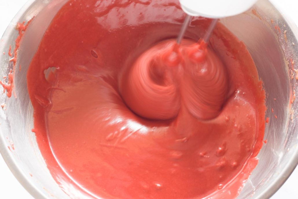 Beating red food coloring into eggless cake batter with a hand mixer.