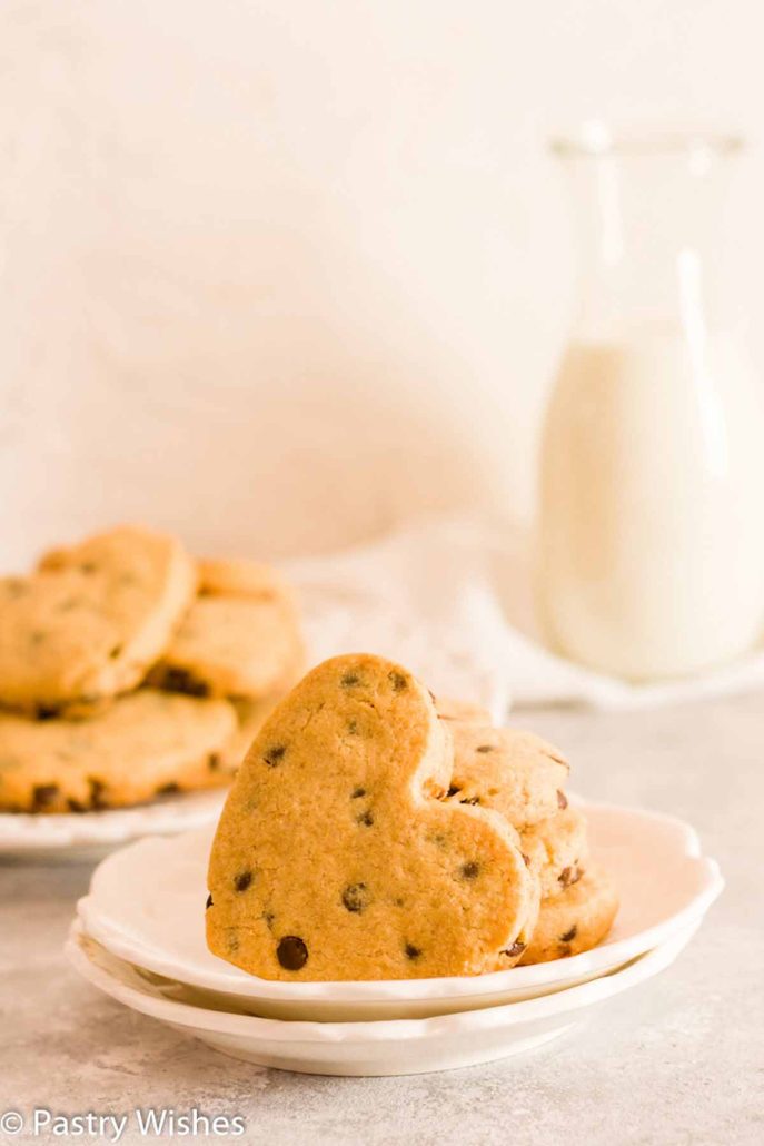 Heart shaped chocolate chip cookies on a white plate with stacked cookies and milk in the background.