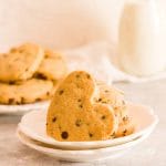 Heart shaped chocolate chip cookies on a white plate with stacked cookies and milk in the background.