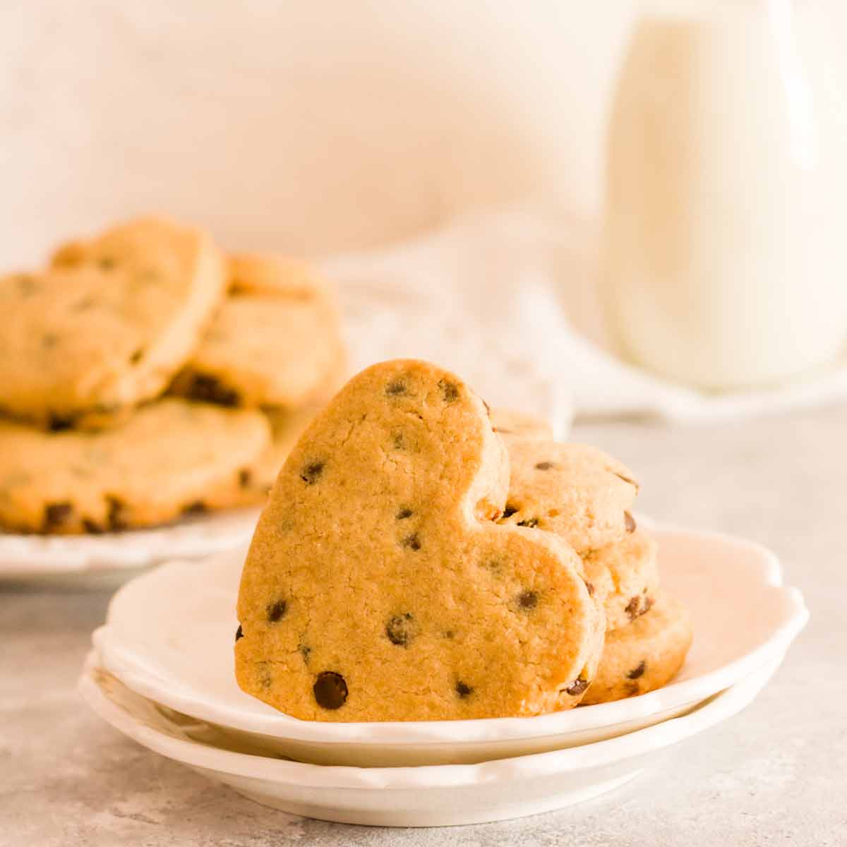 A stack of heart shaped chocolate chip cookies on white plates with milk and cookies in the back.