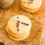 Lemon lavender cookies stacked on each other with lavender flowers on top on a wooden table.