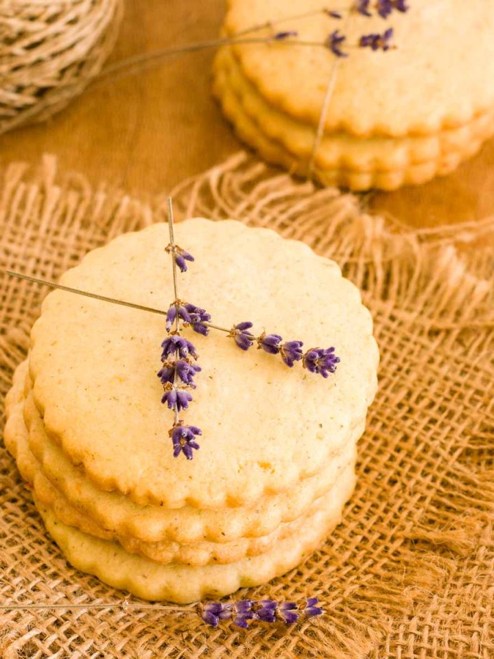 Lemon lavender cookies stacked on each other with lavender flowers on top on a wooden table.