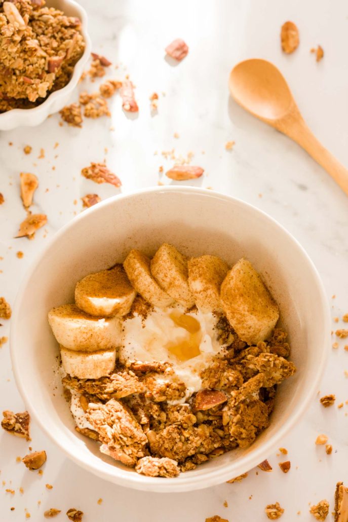 A white bowl filled with Greek yogurt and topped with homemade granola clusters, banana slices, cinnamon and honey on a white surface.