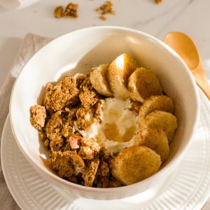 A white bowl filled with Greek yogurt and topped with homemade granola clusters, banana slices, cinnamon and honey.