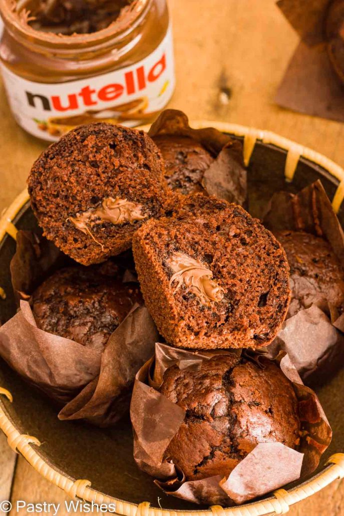 A nutella muffin sliced in half on top of nutella muffins in a wooden bowl next to a jar of nutella on a wooden table.
