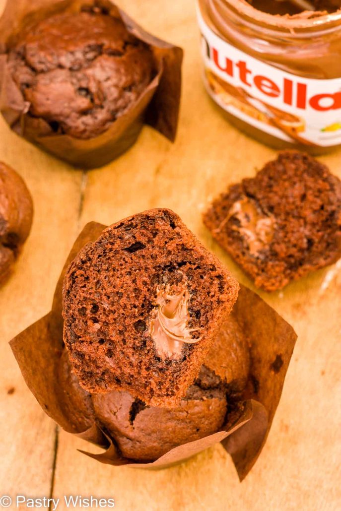 A nutella muffin sliced in half on top of another muffin next to a jar of nutella and a wooden table.