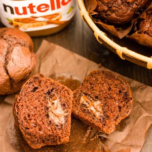 A nutella muffin sliced in half on brown paper next to a jar of nutella and a wooden bowl filled with muffins on a wooden table.
