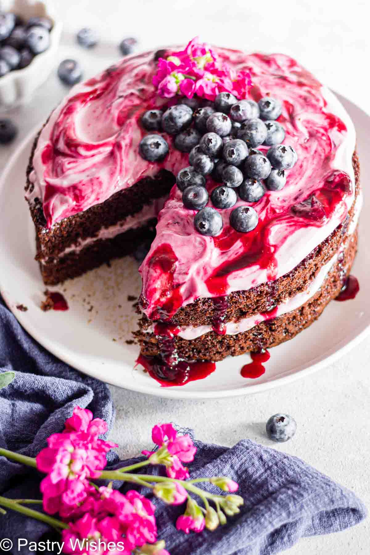 A two layer blueberry chocolate cake on a white plate topped with blueberry cream cheese frosting and blueberries.