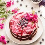 A two layer blueberry chocolate cake on a white plate topped with blueberry cream cheese frosting and blueberries.