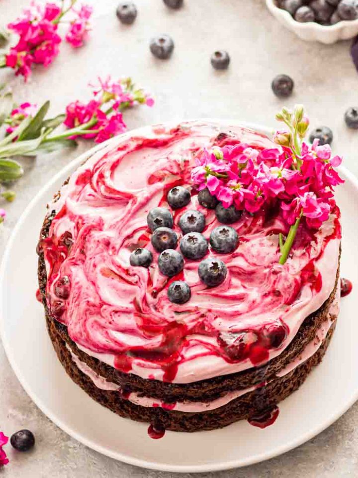 A two layer blueberry chocolate cake on a white plate topped with blueberry cream cheese frosting and blueberries.