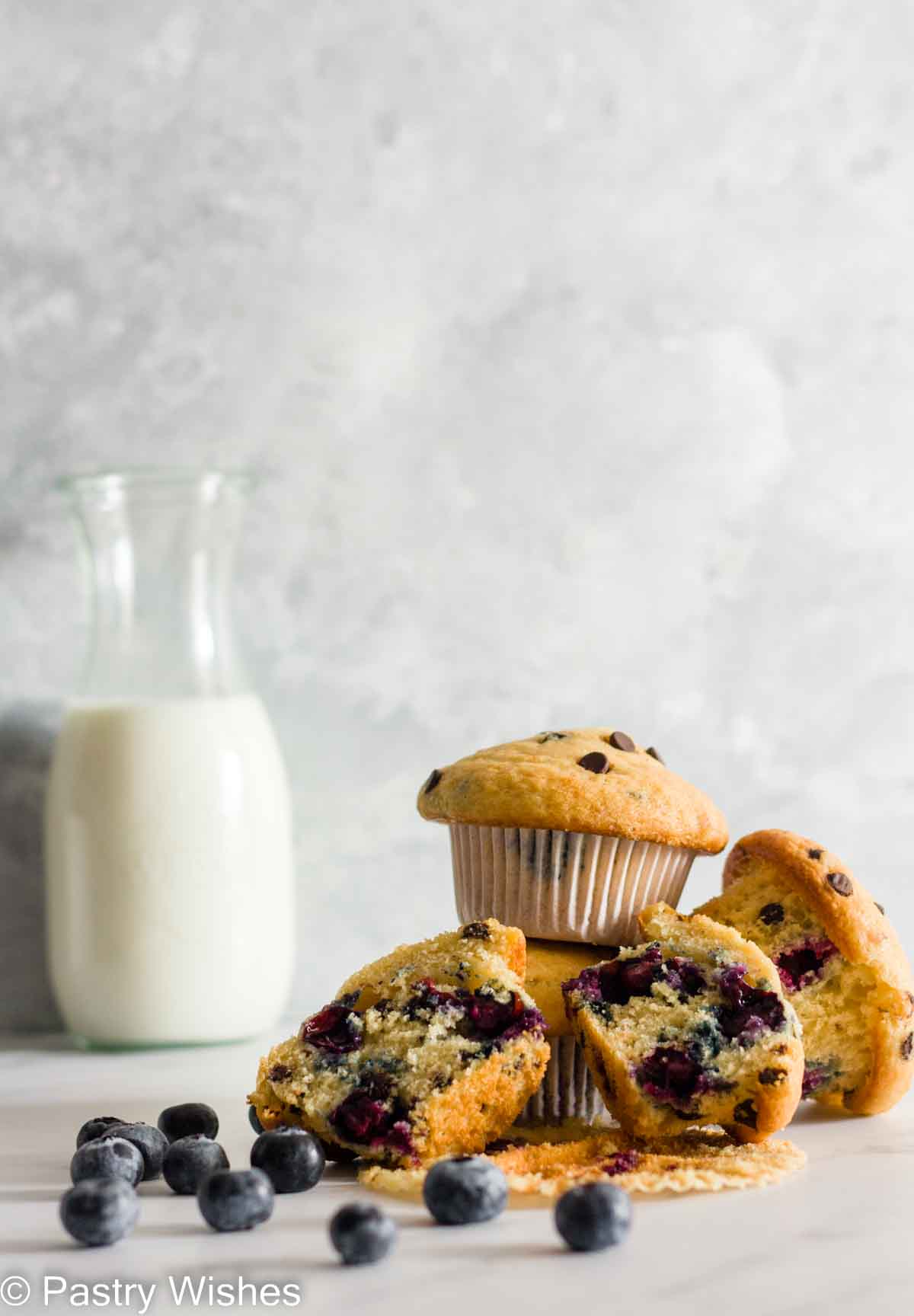 A stack of blueberry chocolate chip muffins on a white surface next to a bottle of milk and blueberries.