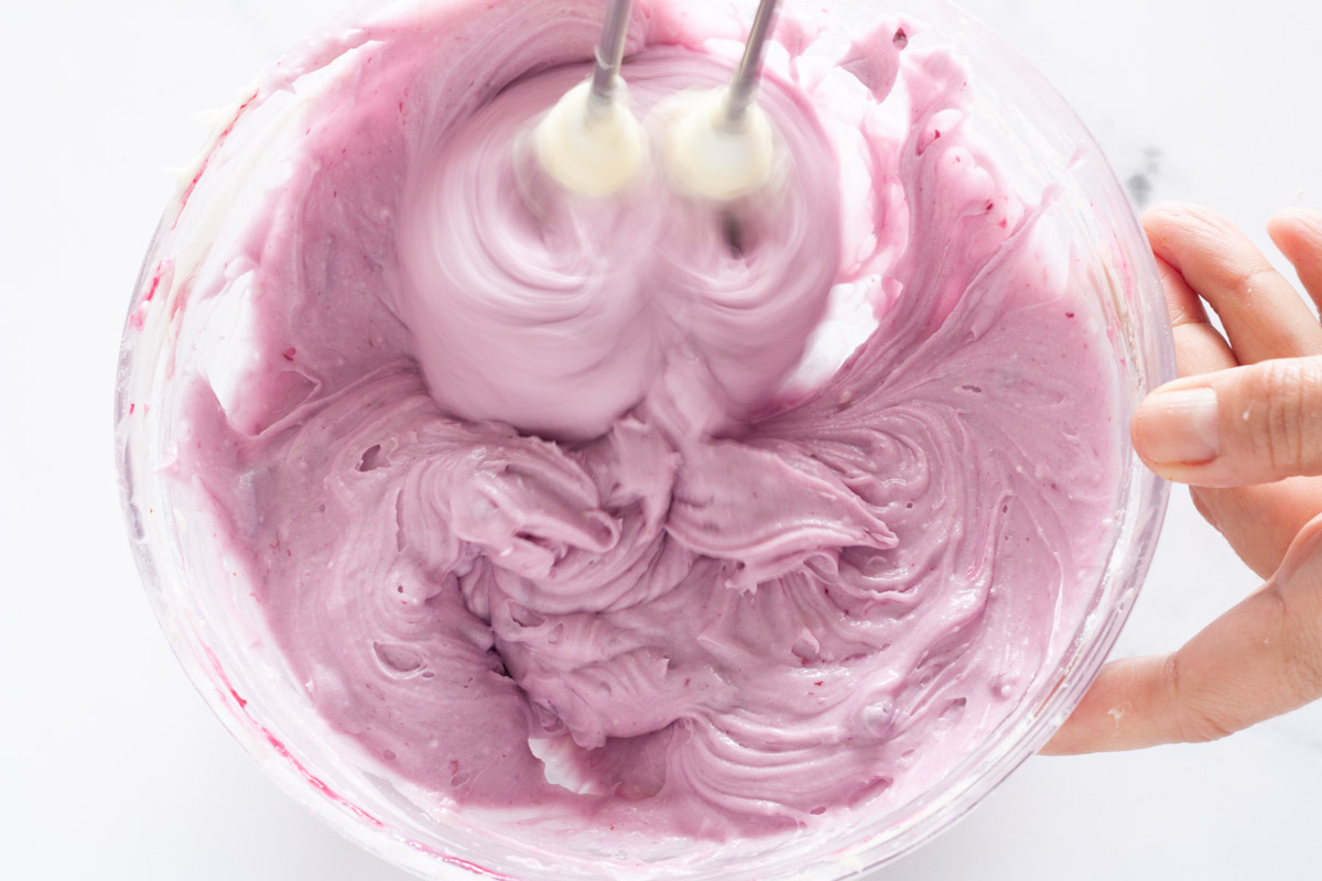 Beating blueberry sauce and cream cheese frosting together with a hand mixer in a glass bowl.