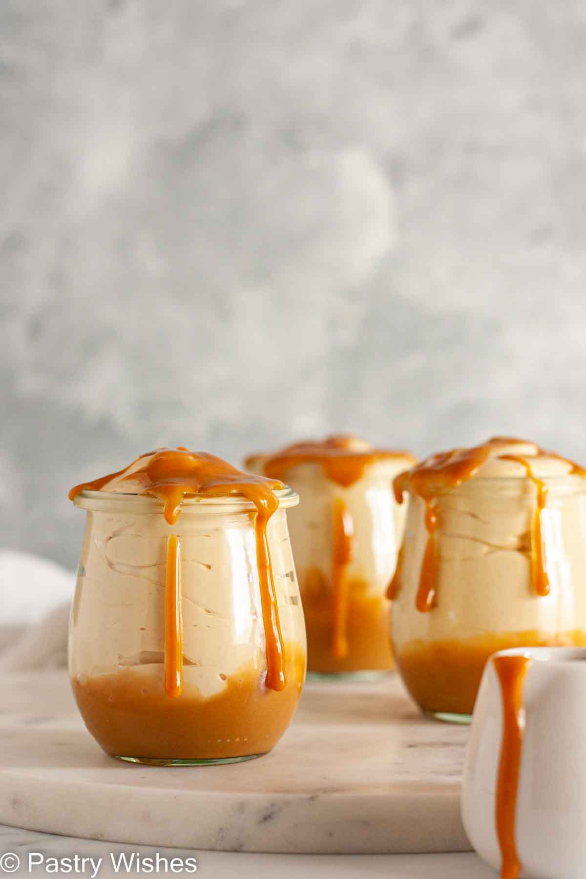 Three serving glasses of Dulce de leche mousse on a white marble surface with dulce de leche dripping on top.