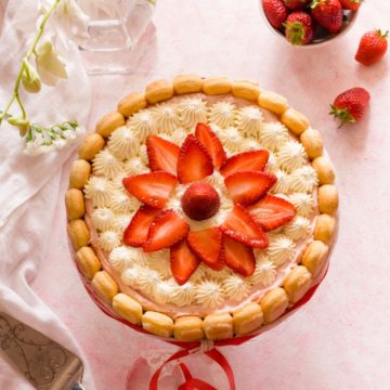 Overhead shot of a strawberry charlotte cake on a pink surface next to strawberries and flowers.