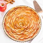 An overhead shot of an apple rose tart with dulce de leche on a white surface next to an apple and cinnamon sticks.