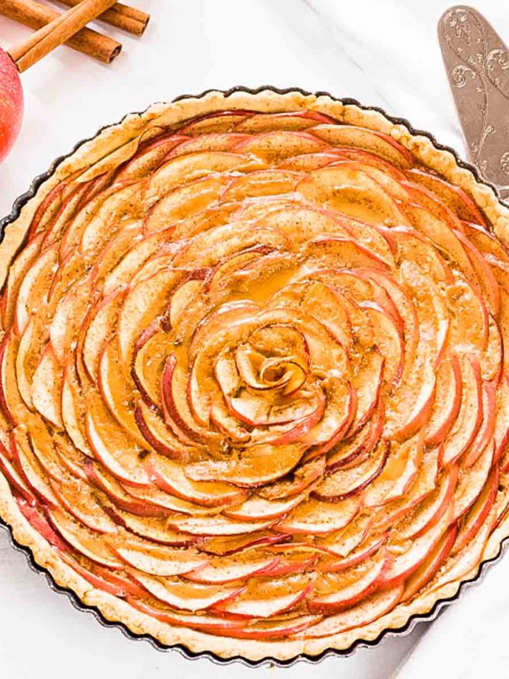 An overhead shot of an apple rose tart with dulce de leche on a white surface next to an apple and cinnamon sticks.